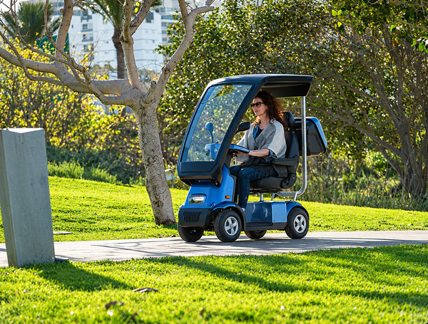 Left Side View Of The AFIKIM Afiscooter C4 Scooter Shown With Optional Hard Top Canopy In The Blue Color Seen In An Outdoor Daytime Park Setting With Trees In The Background