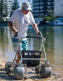 Man Pushing The Wheeleez All-Terrain Beach Rollator Front View With Polyurethane Wheels, Foam Handles, And A Mesh Storage Compartment, Displayed On A Sandy Beach.