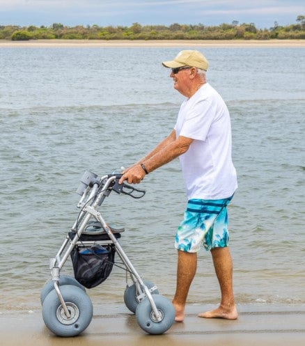 Man Pushing The Wheeleez All-Terrain Beach Rollator Side View With Polyurethane Wheels, Foam Handles, And A Mesh Storage Compartment, Displayed On A Sandy Beach.