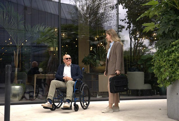 Man Sitting In The Strongback 24 +AB Ultra-Lightweight Wheelchair Conversing With A Woman In A City Setting 