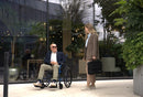 Man Sitting In The Strongback 24 +AB Ultra-Lightweight Wheelchair Conversing With A Woman In A City Setting 
