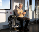 Man Sitting On A Train Using A Smartphone With The Paiseec L5 Mobility Scooter In The Folded Position In The Silver Color Next To Him 