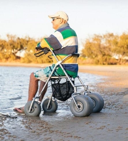 Man Sitting On The Wheeleez All-Terrain Beach Rollator Side View With Polyurethane Wheels, Foam Handles, And A Mesh Storage Compartment, Displayed On A Sandy Beach.