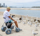 Man Sitting On The Wheeleez All-Terrain Beach Rollator Side View With Polyurethane Wheels, Foam Handles, And A Mesh Storage Compartment, Displayed On A Sandy Beach.