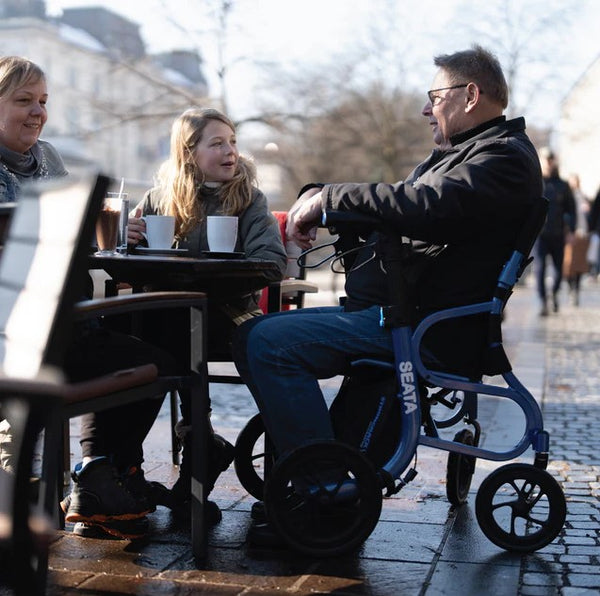 People Sitting At An Outdoor Cafe With A Person Using The Strongback Mobility SEATA Rollator