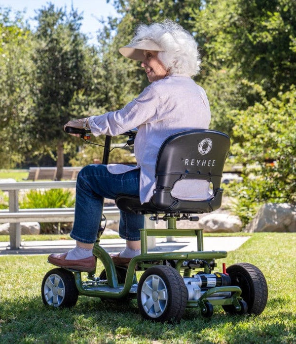 Rear Left Side View Of The Reyhee EXO Electric Mobility Scooter Shown With The Green Frame And An Elderly Woman Riding In An Outdoor Setting During The Day