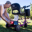 Rear Right Side View Of An Elderly Woman Attaching The Removable Battery Into The Reyhee Triad Compact Folding Electric Mobility Scooter With The Violet Colored Frame In A Daytime Outdoor Setting