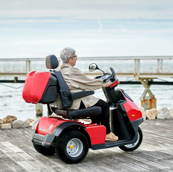 Rear Right Side View Of The AFIKIM Afiscooter S3 Scooter In The Red Color With Optional Rear Lock Box Shown With An Elderly Woman Riding On A Pier With The Ocean In The Background