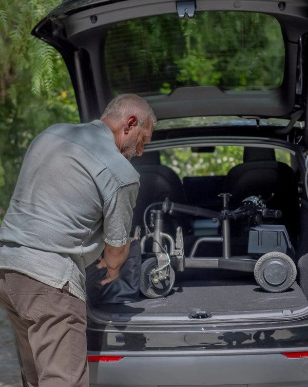 Rear View Of An Elderly Man Loading The Reyhee Triad Compact Folding Electric Mobility Scooter With A Gun Metal Colored Frame Into The Trunk Of His Compact Car In A Daytime Setting