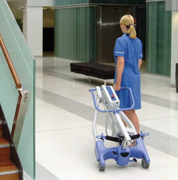 Rear View Of The Joerns Hoyer Advance-E Portable Electric Professional Patient Lift Shown In The Folded Position With A Woman Healthcare Worker Transporting The Lift By Rolling It Using The Rear Casters In A Hospital Setting