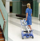 Rear View Of The Joerns Hoyer Advance-E Portable Electric Professional Patient Lift Shown In The Folded Position With A Woman Healthcare Worker Transporting The Lift By Rolling It Using The Rear Casters In A Hospital Setting