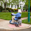 Rear View Of The Journey So Lite Mobility Scooter Shown In Blue Color With Elderly Woman Riding The Scooter In A Neighborhood Setting