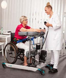 Right Side View Of An Elderly Woman Sitting In A Wheelchair While A Healthcare Worker Assists Her Using The TR Equipment Gate Bure Rise & Go