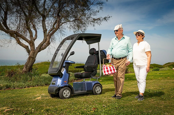 Right Side View Of The AFIKIM Afiscooter C4 Scooter Shown With Optional Hard Top Canopy In The Blue Color With An Elderly Couple Seen Walking On A Grass Field With A Picnic Box On A Sunny Day