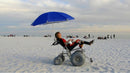 Right Side View Of The Debug Mobility All-Terrain Beach Wheelchair Shown With The Tilt In Space Model In A Beach Setting With A Woman Sitting In The Chair Under An Umbrella
