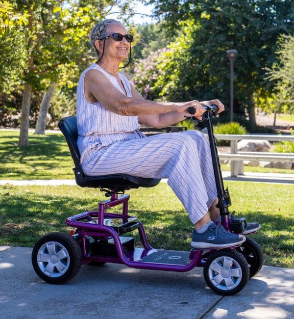 Right Side View Of The Reyhee EXO Electric Mobility Scooter With The Purple Frame And An Elderly Woman Riding In An Outdoor Setting During The Day