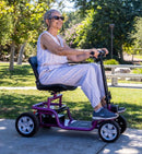 Right Side View Of The Reyhee EXO Electric Mobility Scooter With The Purple Frame And An Elderly Woman Riding In An Outdoor Setting During The Day