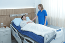 Right Side View Of The Wellell Domus Auto Pressure Redistribution Mattress System With Quilted Cover Shown On An Adjustable Hospital Bed In A Hospital Room Setting With Patient Lying In Bed With Healthcare Worker Looking Over Him With Her Hand Placed On His Shoulder
