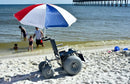 Side View Of The Debug Mobility All-Terrain Beach Wheelchair With An Umbrella Shown In A Beach Setting With The Ocean In The Background