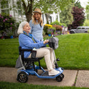 Side View Of The Journey So Lite Mobility Scooter Shown In Blue Color Riding On Sidewalk With Elderly Woman And Middle Aged Woman