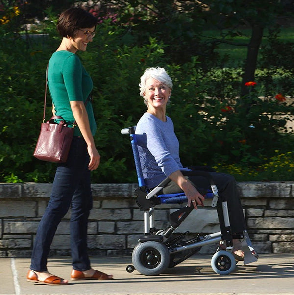 Side View Of The Journey Zinger Electric Wheelchair Shown In Blue Color With Elderly Woman In Chair With Another Woman Standing Beside Her