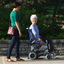 Side View Of The Journey Zinger Electric Wheelchair Shown In Blue Color With Elderly Woman In Chair With Another Woman Standing Beside Her