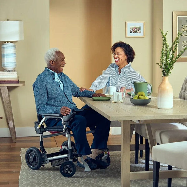 Side View Of The Journey Zoomer Electric Wheelchair Shown In Black Color With Elderly Man Sitting In The Chair With Woman Sitting Across From Him
