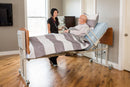Side View Of The Med Mizer EX8000 Bariatric Adjustable Bed Shown With Two Pivoting Assist Bars And Cherry Board Finish With Elderly Man Lying Down And Nurse Smiling At Him In A Homecare Setting