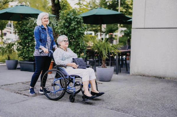 Two Women, One In The Strongback 24 Flip Back Arms Ultra-Lightweight Wheelchair, Walking Outdoors On A Sidewalk With Greenery And Building In The Background