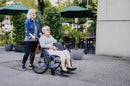 Two Women, One In The Strongback 24 Flip Back Arms Ultra-Lightweight Wheelchair, Walking Outdoors On A Sidewalk With Greenery And Building In The Background