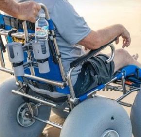 Wheeleez Sandcruiser All-Terrain Beach Wheelchair Rear View In Mediterranean Blue Color With Large Balloon Wheels And RoboCup Holder, Positioned On A Sandy Beach With Man Sitting In The Wheelchair While A Man Stands Behind The Wheelchair Holding The Handles 