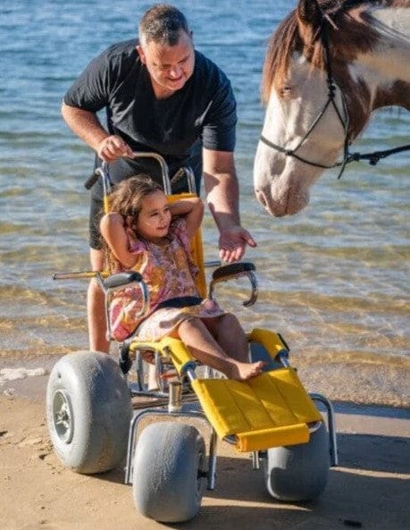 Wheeleez Sandpiper All-Terrain Beach Wheelchair Front View Yellow And Silver Color With Large Wheels Designed For Children And Small Adults On A Sandy Beach With A Young Girl Sitting With Her Father Standing Behind The Wheelchair Petting A Horse And The Ocean In The Background.