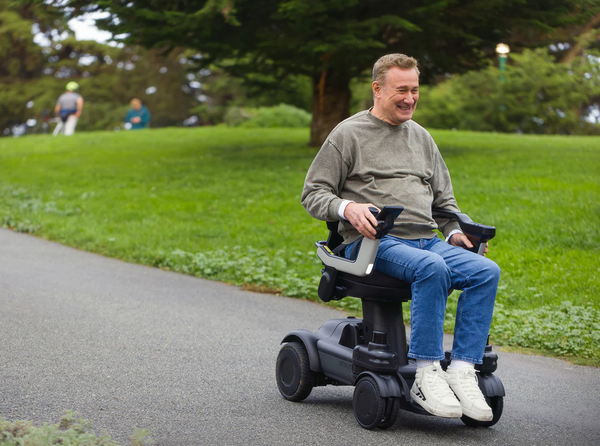 Wide Angle Shot Of A Man Smiling While Riding The STRUTT ev1 Smart Electric Mobility Vehicle Down A Paved Road Shown In An Outdoor Park Setting