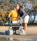 Woman And Child Standing Next To The Wheeleez All-Terrain Beach Rollator Front View With Polyurethane Wheels, Foam Handles, And A Mesh Storage Compartment, Displayed On A Sandy Beach.