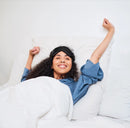 Woman Lying In Bed With A Rize Home Silk Sleep Mask On While Stretching And Smiling