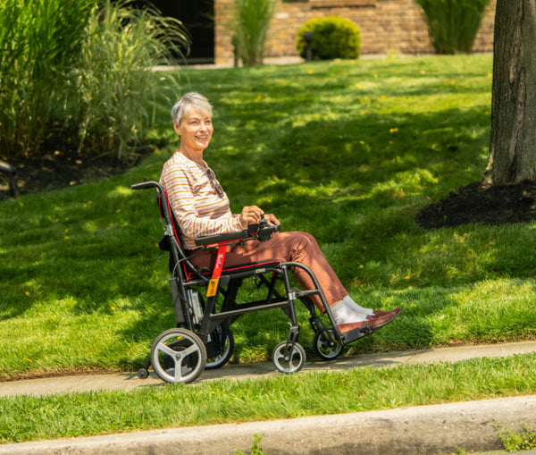 Woman Sitting In The Feather Electric Wheelchair On A Sidewalk With A Yard In The Background