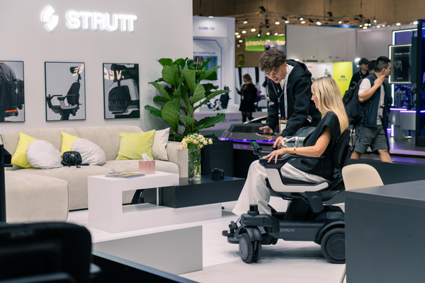 Woman Sitting In The STRUTT ev1 Smart Electric Mobility Vehicle With A Young Man Explaining The Features Of The Device Shown In A Convention Center Setting