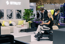 Woman Sitting In The STRUTT ev1 Smart Electric Mobility Vehicle With A Young Man Explaining The Features Of The Device Shown In A Convention Center Setting