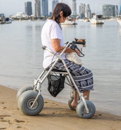 Woman Sitting With Dog On The Wheeleez All-Terrain Beach Rollator Side View With Polyurethane Wheels, Foam Handles, And A Mesh Storage Compartment, Displayed On A Sandy Beach.