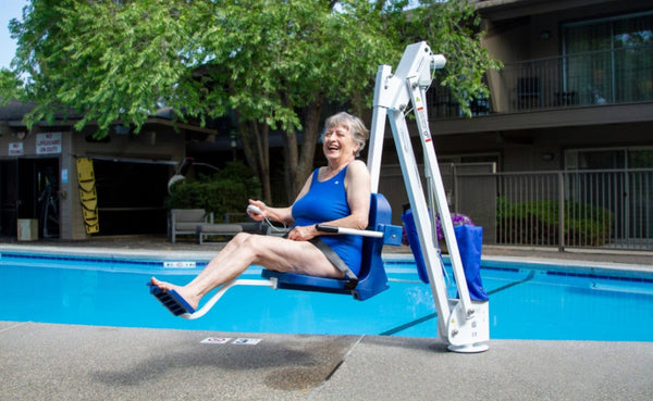Woman Smiling While Using The Aqua Creek Mighty 400 Pool Lift Blue And White Color Front View Next To A Swimming Pool With Trees And Building In The Background.