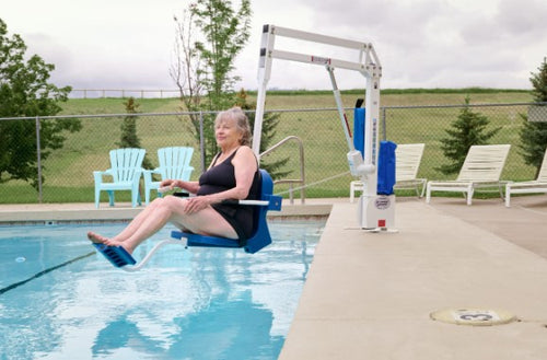 Woman Using The Aqua Creek Scout Excel Pool Lift Blue Color To Enter A Pool With Chairs And Greenery In The Background.