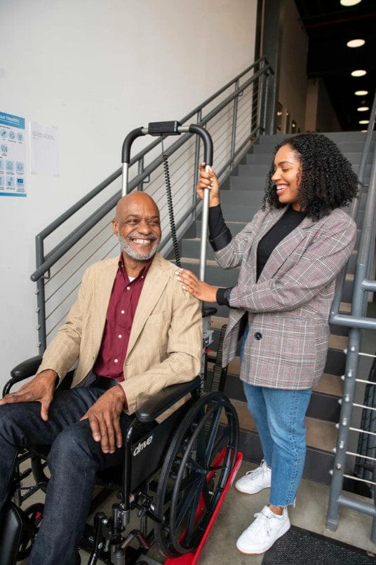 A Woman Smiling While Standing Next To A Smiling Elderly Man Sitting In A Wheelchair Inside The Atlas Stair Climbing Wheelchair Lift Shown With A Flight Of Stairs In The Background