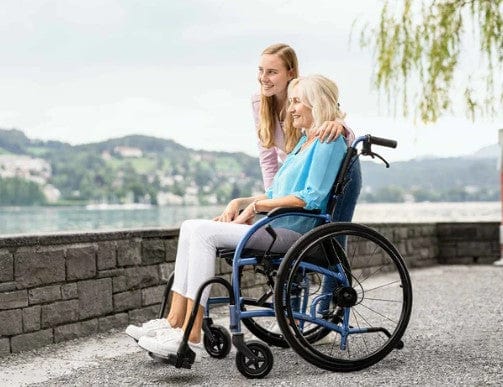 Elderly Woman Sitting In The Strongback 24 +AB Ultra-Lightweight Wheelchair With Younger Woman Standing Beside Her In An Outdoor Setting