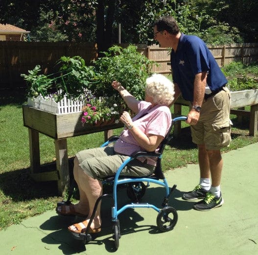 Elderly Woman Sitting In The Strongback Excursion 8 Lightweight Transport Chair With A Man Holding The Handles Behind Her Shown In An Outdoor Setting