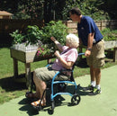 Elderly Woman Sitting In The Strongback Excursion 8 Lightweight Transport Chair With A Man Holding The Handles Behind Her Shown In An Outdoor Setting