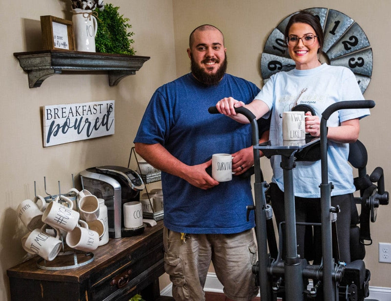 Front View Of The EasyStand Glider Large Active Standing Frame Shown With Smiling Woman Using The Device In A Home Setting With Man Standing Next Her