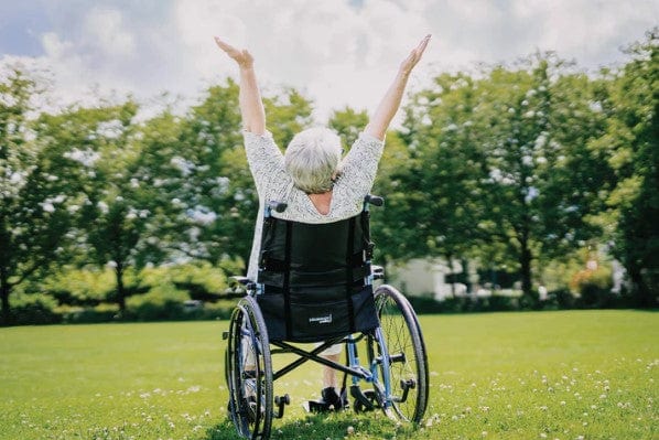 Rear View Of Elderly Woman Raising Her Hands In A Joyous Moment While Sitting In An Outdoor Setting On The Strongback 24 Flip Back Arms Ultra-Lightweight Wheelchair