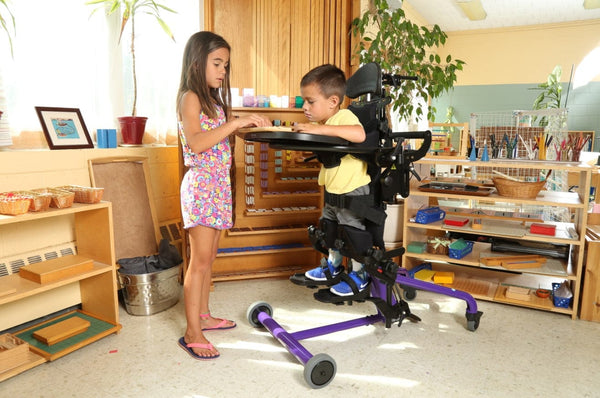 Side View Of The EasyStand Bantam Extra Small Standing Frame Shown In Purple Color With Child Using The Device And Young Girl Standing Next To Him