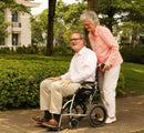 Side View Of The Karman Ergo Lite Transport Wheelchair Shown With An Elderly Man Sitting In The Chair And Elderly Woman Pushing The Chair In A Neighborhood Setting