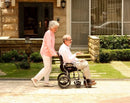 Side View Of The Karman Ergo Lite Transport Wheelchair Shown With An Elderly Man Sitting In The Chair And Elderly Woman Pushing The Chair In A Park Setting